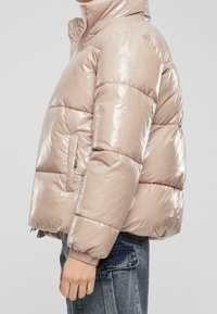 Wypełniona kurtka puchowa o krótkim kroju w połyskującym, jasnobeżowym kolorze. Posiada wysoki kołnierz, zamek błyskawiczny oraz elastyczne mankiety. Widoczne są dwie boczne kieszenie.