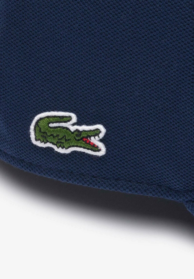 Zielone haftowane logo krokodyla z czerwonymi detalami na ciemnoniebieskiej teksturowanej tkaninie, prawdopodobnie element odzieży.