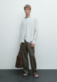 Chemise blanche en lin à boutons sur un t-shirt gris, pantalon vert olive, tongs noires et un sac en cuir marron. Design simple et décontracté.