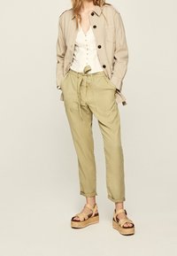 Giacca di cotone beige, camicia bianca con bottoni, pantaloni verdi oliva con vita regolabile, e sandali beige con zeppa e cinghie intrecciate.