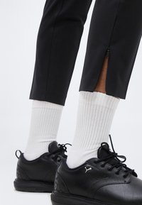 Πόδια που φορούν μαύρο παντελόνι αστραγάλου με φερμουάρ, λευκές ριγωτές κάλτσες και μαύρα παπούτσια Puma με κορδόνια και λογότυπο στο πλάι.