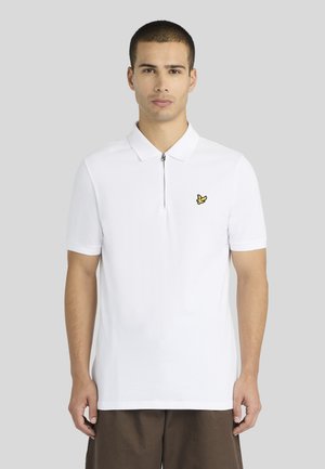 Homme debout, face à l’avant, portant un polo blanc à manches courtes avec une fermeture éclair et un petit logo jaune sur la poitrine.