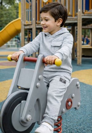 Jeune garçon en sweat à capuche gris et pantalon souriant en position assise et se balançant sur un ressort gris en forme de moto dans une aire de jeux.