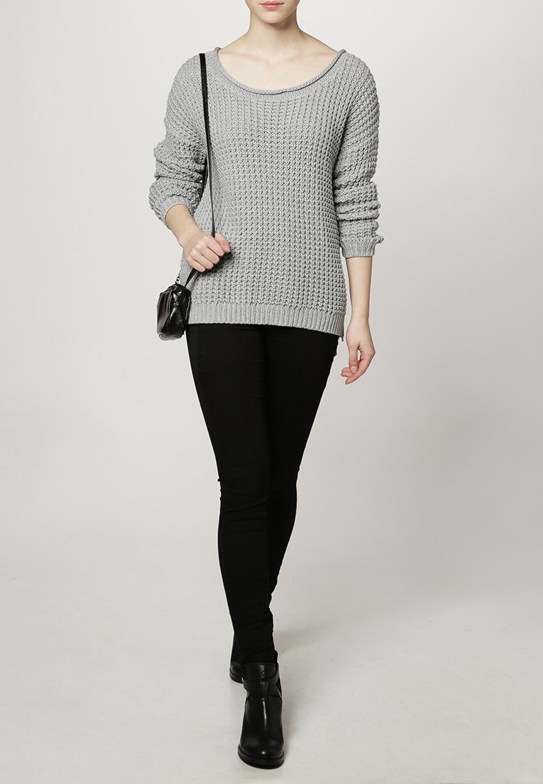 Femme portant un pull en maille gris, un pantalon skinny noir, des bottines noires, et un petit sac bandoulière noir, marchant vers l'avant.