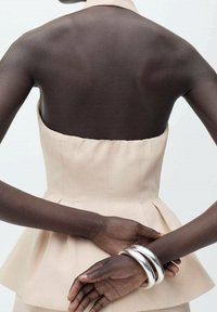 Peplum-Top aus hellbeigem Stoff mit schulterfreiem Design. Enthält silberne Armreifen an den Handgelenken, von hinten gezeigt. Glatte Textur, maßgeschneiderte Passform.