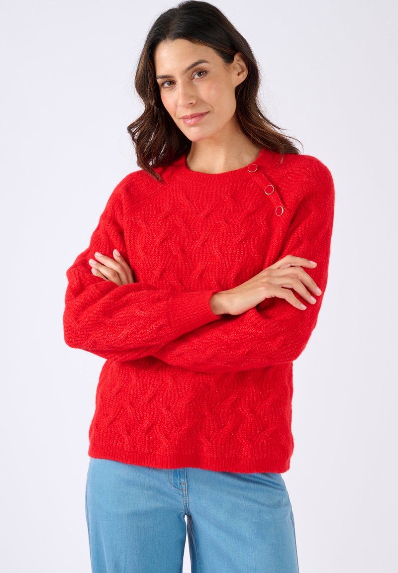 Donna con capelli scuri che indossa un maglione rosso brillante a trecce con dettagli in bottone sulla spalla e jeans azzurri chiari, su sfondo bianco.