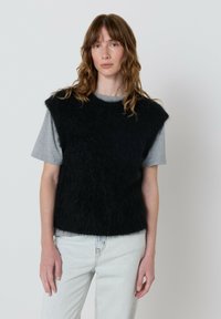 Jauna moteris su banguotais rudais plaukais, vilkinti juodą pūkuotą be rankovių megztinį ant pilkos marškinėlių ir šviesiai mėlynų džinsų, stovi prieš paprastą fono.