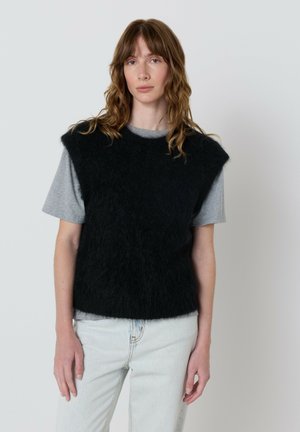 Mladá žena s vlnitými hnedými vlasmi, oblečená v čiernom chlpatom svetri bez rukávov cez sivé tričko a svetlomodré džínsy, stojí na neutrálnom pozadí.