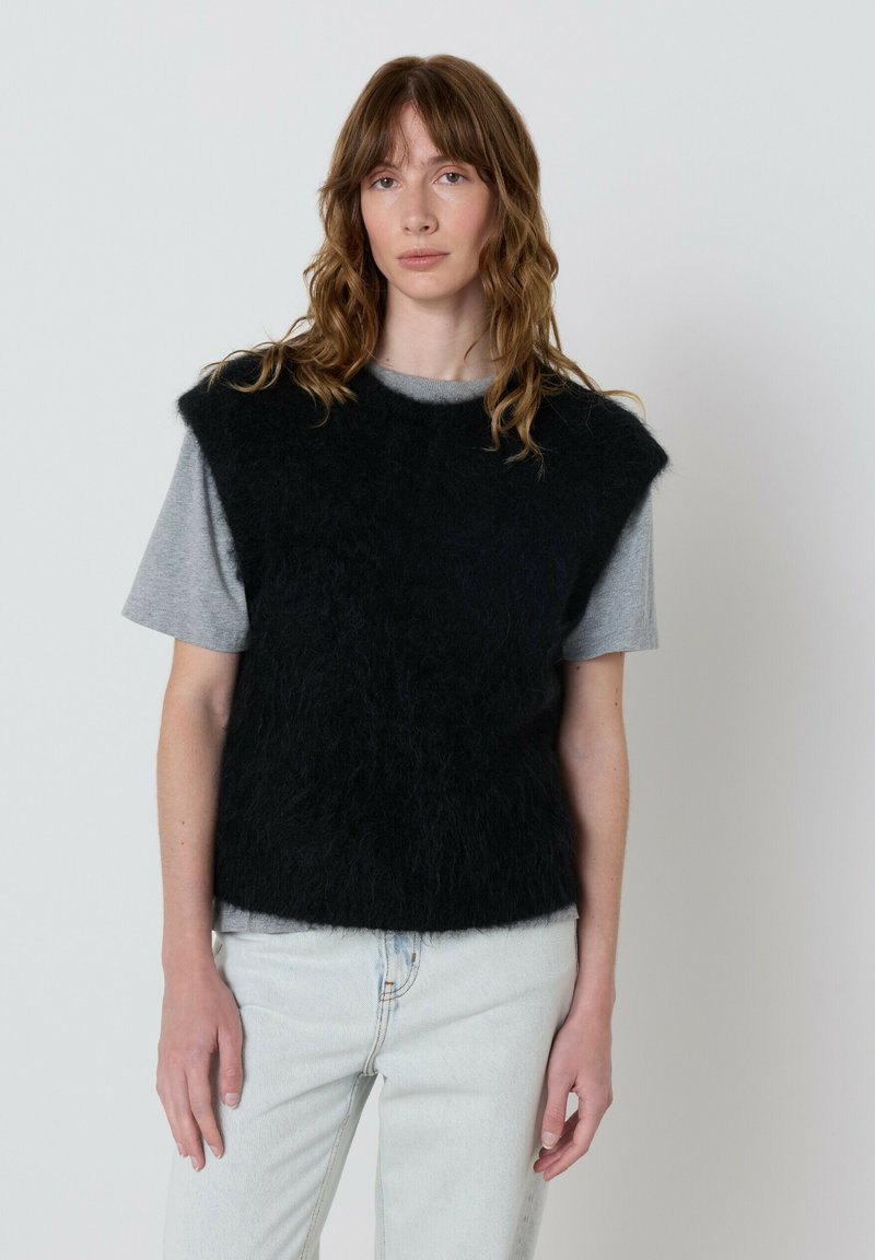 Jauna moteris su banguotais rudais plaukais, vilkinti juodą pūkuotą be rankovių megztinį ant pilkos marškinėlių ir šviesiai mėlynų džinsų, stovi prieš paprastą fono.