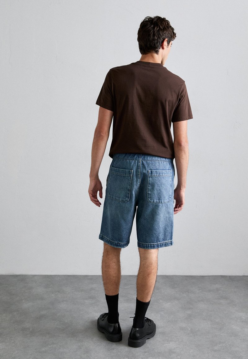 T-shirt in cotone marrone abbinato a pantaloni shorts in denim azzurro chiaro con una vestibilità rilassata, due tasche posteriori e orli arrotolati. Calzini e scarpe neri.