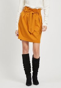 Jupe orange à ceinture avec des plis, en tissu structuré. Portée avec des bottes noires molles jusqu'aux genoux. Un haut blanc simple complète la tenue.