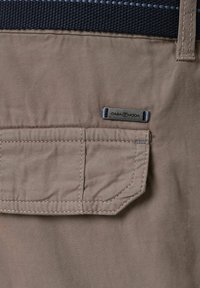 Brauner Stoff mit strukturierter Oberfläche, ausgestattet mit einer Klapptasche und einem Metallschild. Der Taillengürtel ist dunkelblau mit einem gewebten Muster.