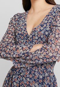 Vestido floral con un profundo escote en V, mangas largas y transparentes, y un corsé ajustado. El fondo azul marino está adornado con pequeñas flores rosas y azules.