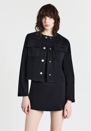 Black cropped jacket in soft fabric, featuring two front pockets, snap buttons, and long sleeves. Worn with a fitted black skirt.