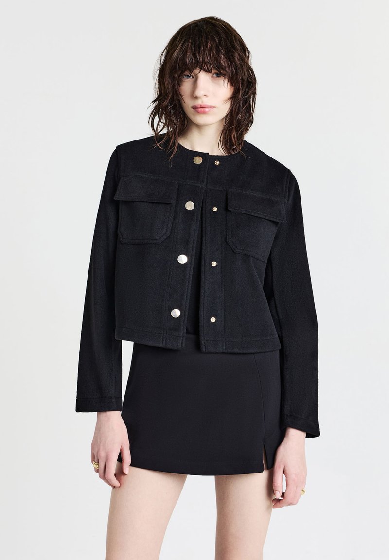 Giacca nera corta in tessuto morbido, con due tasche frontali, bottoni a pressione e maniche lunghe. Indossata con una gonna nera aderente.
