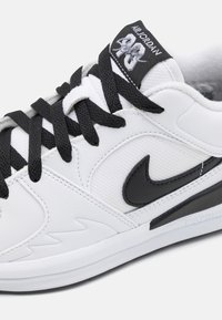 Fehér sneaker fekete fűzőkkel és akcentusokkal, textúrált felsőrésszel, kiemelkedő fekete Nike logóval és párnázott nyelvvel, amelyen a márkajelzés található.