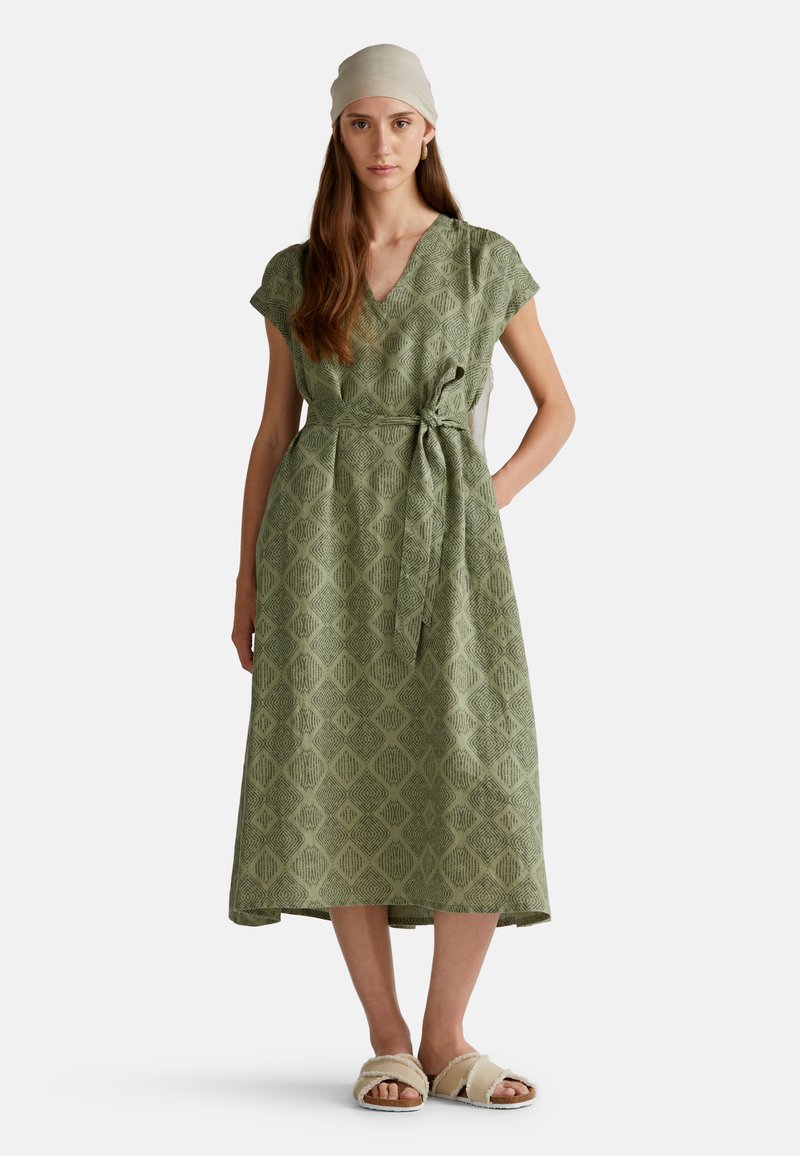 Robe midi à motifs verts avec des manches courtes, un design enroulé et une taille nouée. Dotée d'un motif losange et de poches latérales, associée à des sandales beiges.
