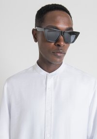 Occhiali da sole neri e angolari con lenti scure; indossati con una camicia bianca con bottoni, caratterizzata da un colletto rialzato e un tessuto texture.