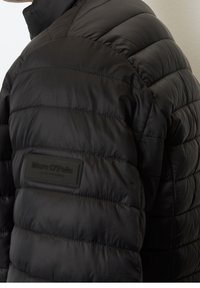 Sort pufferjakke med quiltet design og høj krave. Har et tekstureret logo-patch på venstre side. Holdbart, let materiale.