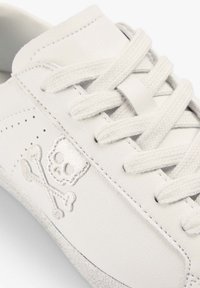 Baskets blanches avec lacets en tissu et un emblème cousu de tête de mort et os croisés sur le côté près des œillets.