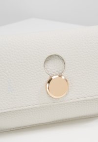 Biała skórzana kopertówka o fakturze z okrągłym, złotym zapięciem zatrzaskowym oraz wycięciem. Gładkie wykończenie z widocznym szwem wzdłuż krawędzi.
