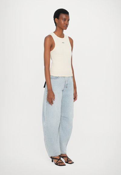 Crème gekleed rib tanktop met een ronde halslijn, gecombineerd met lichtblauwe wijde jeans en zwarte strappy hakken. Achterzijde met strikdetail zichtbaar.