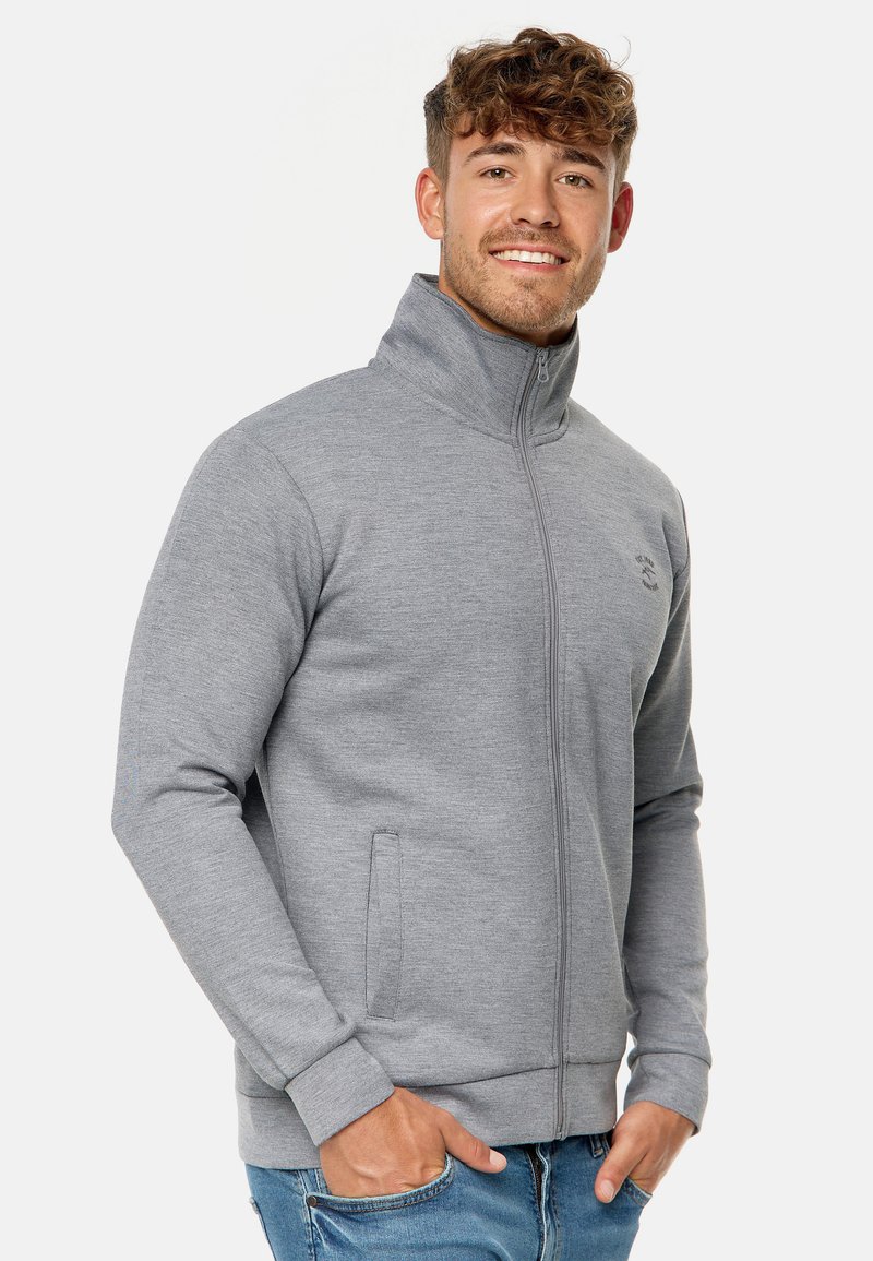 Graue Zip-Jacke mit hohem Kragen, gerippten Bündchen und seitlichen Taschen. Weiche Textur mit dezentem Logo-Detail auf der Brust.