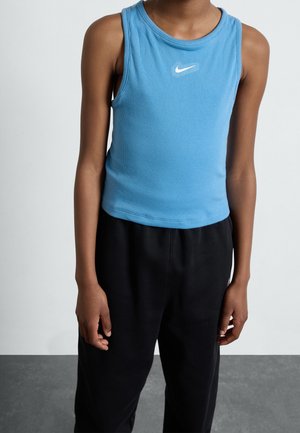 Dziecko ubrane w niebieską bezrękawnik Nike i luźne czarne spodnie stoi na jednolitym tle.