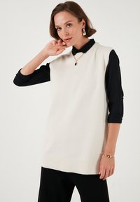 Sleeveless cream knit tunic with ribbed hem, layered over black shirt with rolled sleeves. Gold chain necklace with geometric pendant.