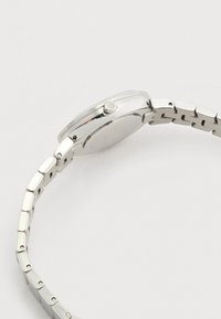 Ασημένιο μεταλλικό ρολόι χειρός με συνδεδεμένο λουράκι, που δείχνει την πλευρική όψη εστιασμένη στο κορώνα και το πίσω μέρος της θήκης, σε λευκό φόντο.