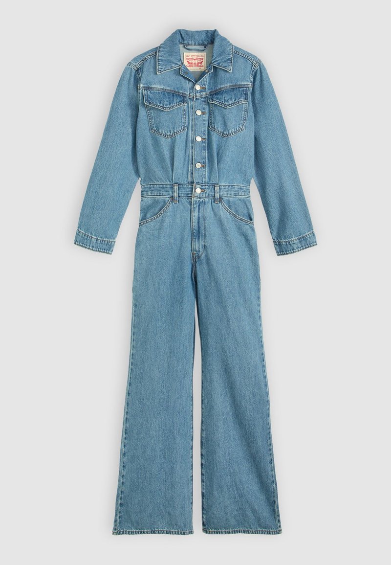 Combinaison en denim bleu clair à manches longues, avec une fermeture à boutons sur le devant, deux poches poitrine, et une coupe jambes larges. Présente des surpiqûres et un col classique.