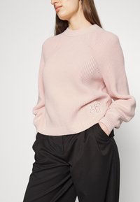 Femme portant un pull en maille rose clair avec un détail de logo et un pantalon taille haute noir, les mains dans les poches, sur un fond uni.