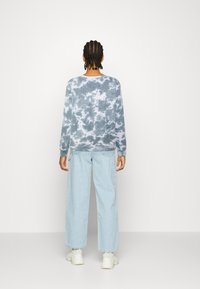 Světle modrý tie-dye svetr s dlouhými rukávy, doplněný světle modrými širokými džínami. Model má na sobě bílé robustní tenisky. Pozadí je jednobarevné bílé.