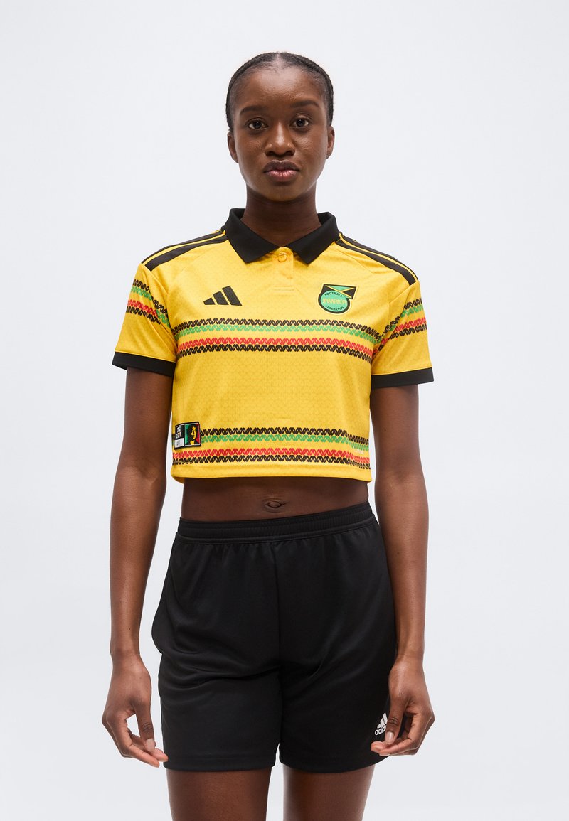 Νέα γυναίκα που φοράει ένα κίτρινο κοντό ποδοσφαιρικό φανελάκι με πράσινες, κόκκινες και μαύρες ρίγες και μαύρο σορτς, στέκεται μπροστά σε έναν απλό φόντο.