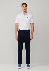 Camisa polo blanca con un bolsillo en el pecho, combinada con pantalones ajustados azul marino y zapatillas azules, con una textura suave y líneas limpias.