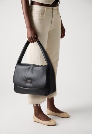 Borsa a tracolla in pelle nera con una texture liscia, una singola tracolla e una patta frontale con un gancio in argento, tenuta da una persona.