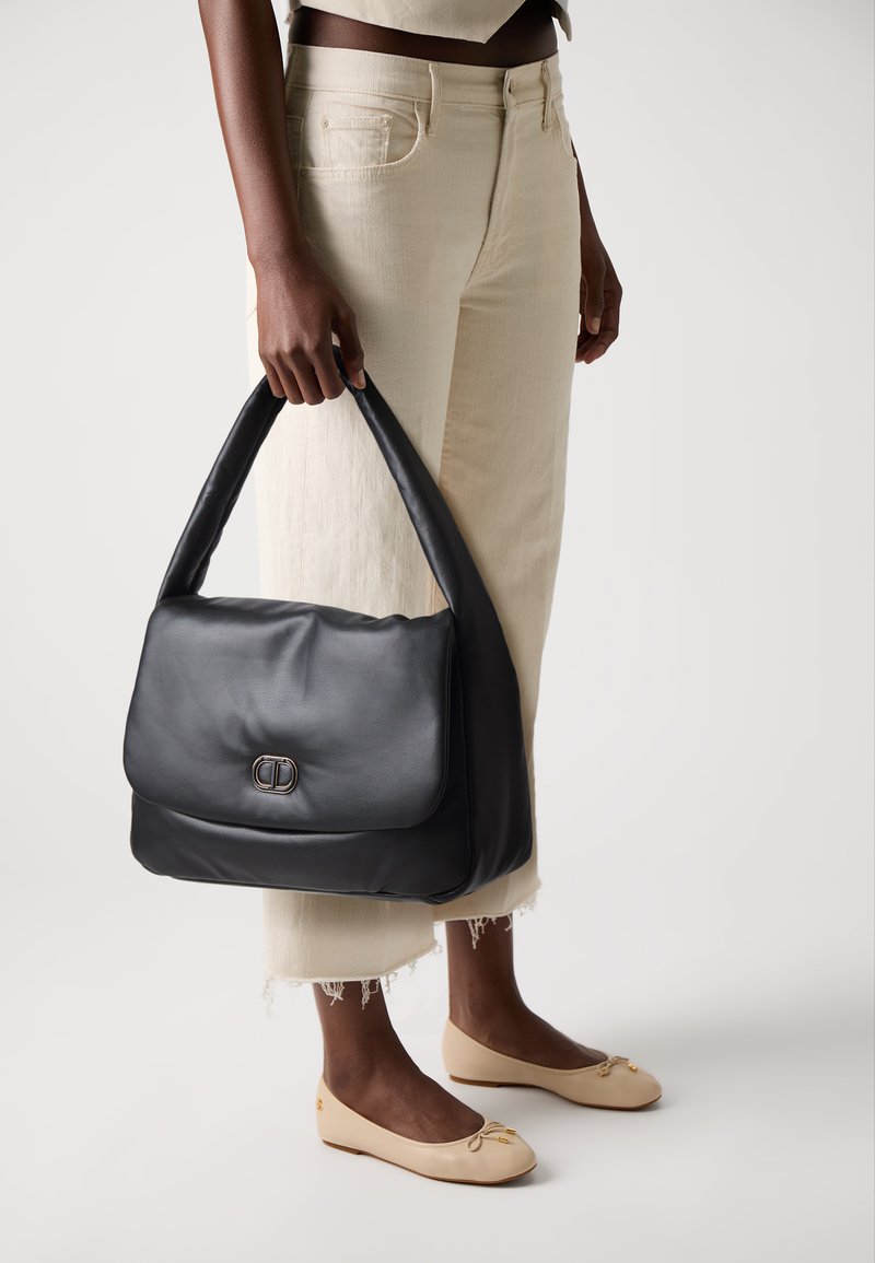 Borsa a tracolla in pelle nera con una texture liscia, una singola tracolla e una patta frontale con un gancio in argento, tenuta da una persona.