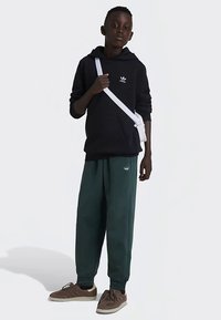 Μαύρη φόρμα με λευκό λογότυπο, πράσινες φόρμες, και καφέ αθλητικά παπούτσια. Το μοντέλο κρατάει μια λευκή τσάντα χιαστί. Λείος ύφασμα, χαλαρή εφαρμογή.