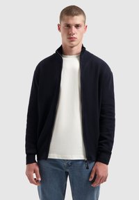 Marineblauwe geribbelde zip-up jas met hoge kraag, gecombineerd met een wit T-shirt en lichtblauwe jeans. Heeft elastische boorden en zijzakken.