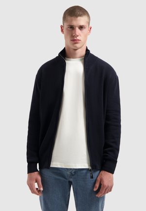 Marineblauwe geribbelde zip-up jas met hoge kraag, gecombineerd met een wit T-shirt en lichtblauwe jeans. Heeft elastische boorden en zijzakken.