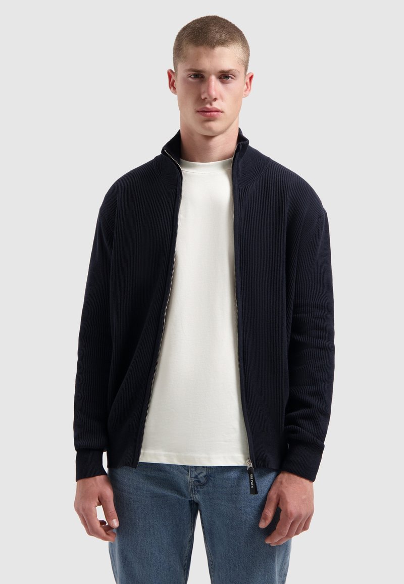 Marineblauwe geribbelde zip-up jas met hoge kraag, gecombineerd met een wit T-shirt en lichtblauwe jeans. Heeft elastische boorden en zijzakken.