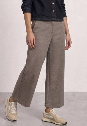 Personne portant un pantalon marron à jambes larges, une veste courte foncée et des baskets beiges, debout sur un sol gris.