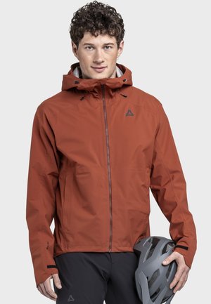 Junger Mann trägt eine rostfarbene Kapuzenjacke und hält einen grauen Fahrradhelm in seiner rechten Hand vor einem schlichten Hintergrund.