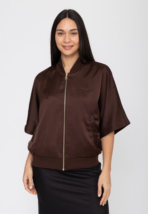 Vrouw met lang donker haar, gekleed in een bruine satijnen jas met korte mouwen en een zwarte rok, glimlachend tegen een neutrale achtergrond.