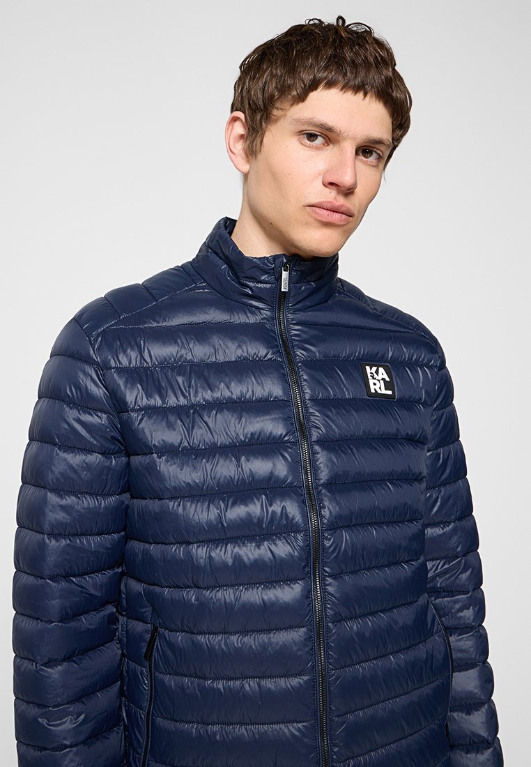Joven con chaqueta acolchada azul marino con cremallera y pequeño logo blanco "KARL" en el pecho, de pie frente a un fondo liso.