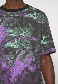 T-shirt à manches courtes noir avec des motifs abstraits en violet et vert menthe. Présente un col rond avec une bordure noire contrastante.