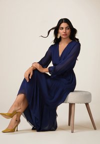 Robe portefeuille bleu marine avec de longues manches en voile et une jupe fluide ; présente un décolleté en V profond et une taille froncée. Assortie à des escarpins jaunes.