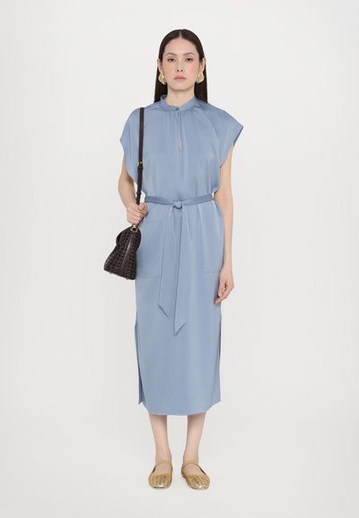 Ανοιχτό μπλε μίντι φόρεμα με κοντά μανίκια, ζώνη στη μέση και δύο μπροστινές τσέπες. Συνδυασμένο με μια μαύρη πλεκτή τσάντα και χρυσά φλατ παπούτσια.