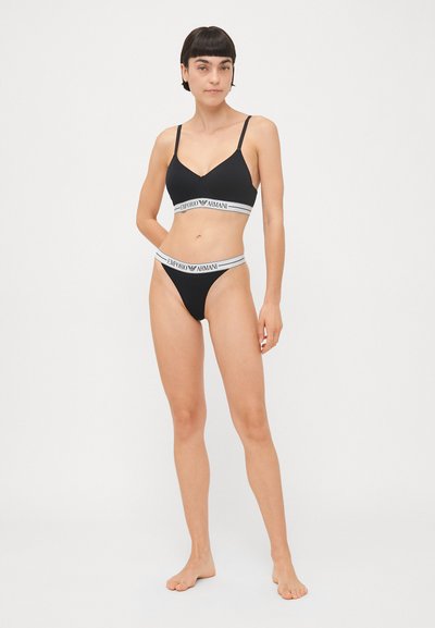 Černý bikinový set s braletou s nastavitelnými ramínky a odpovídajícími tanga kalhotkami. Obě části mají bílé elastické pásky s logem.