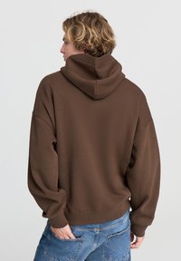 Hoodie oversize marrone con cappuccio regolabile, una texture morbida e spalle scese, abbinato a jeans azzurri chiari.